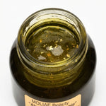 An open dark amber glass jar containing a translucent olive-green body gel with visible gold flakes, featuring a beige label that reads 'MOLIAE Beauty "Ahya Gold Kingship"'.